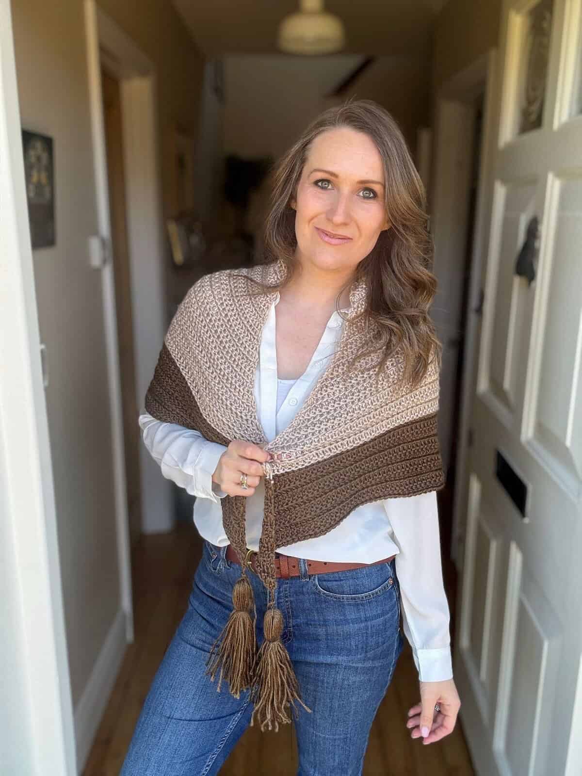A woman stands in a hallway wearing a white blouse, blue jeans, and a brown crochet boomerang shawl with tassels, looking at the camera.