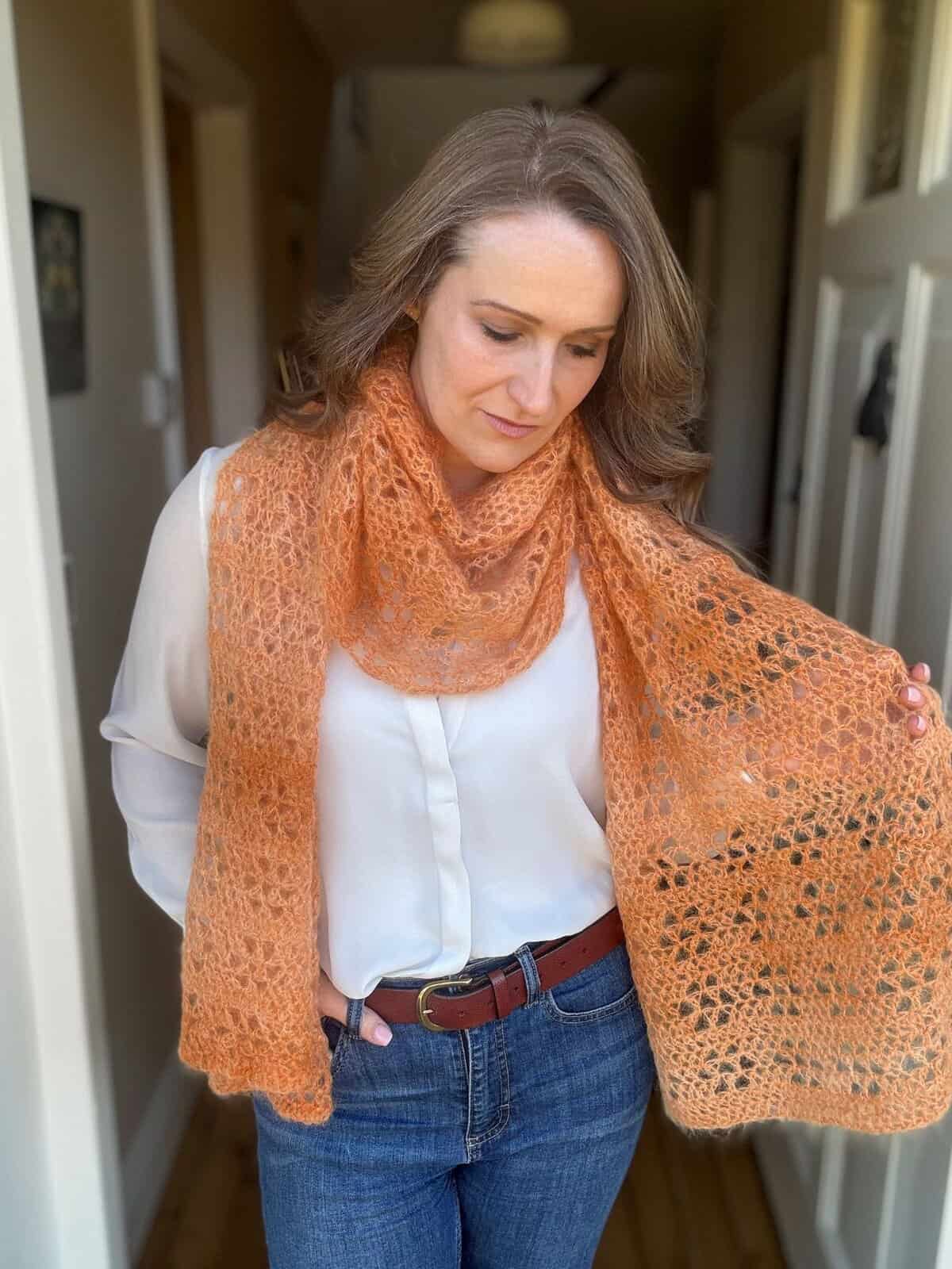 A woman stands indoors wearing a white blouse and blue jeans, draped in a light orange, lacy crochet boomerang shawl.