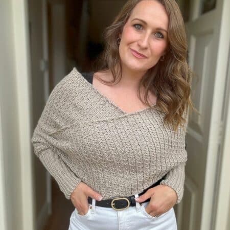 A woman with wavy brown hair stands indoors, wearing a beige off-shoulder crochet sweater scarf and white pants, her hand in her pocket while showcasing a scarf with sleeves crochet pattern draped elegantly over her shoulder.
