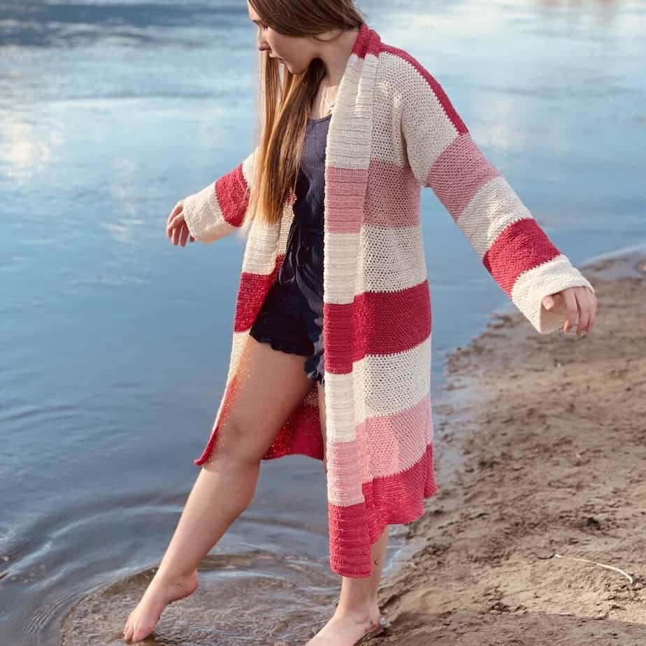 young woman wearing pink and white stripe long sleeved crochet cardigan dipping her toe in the sea