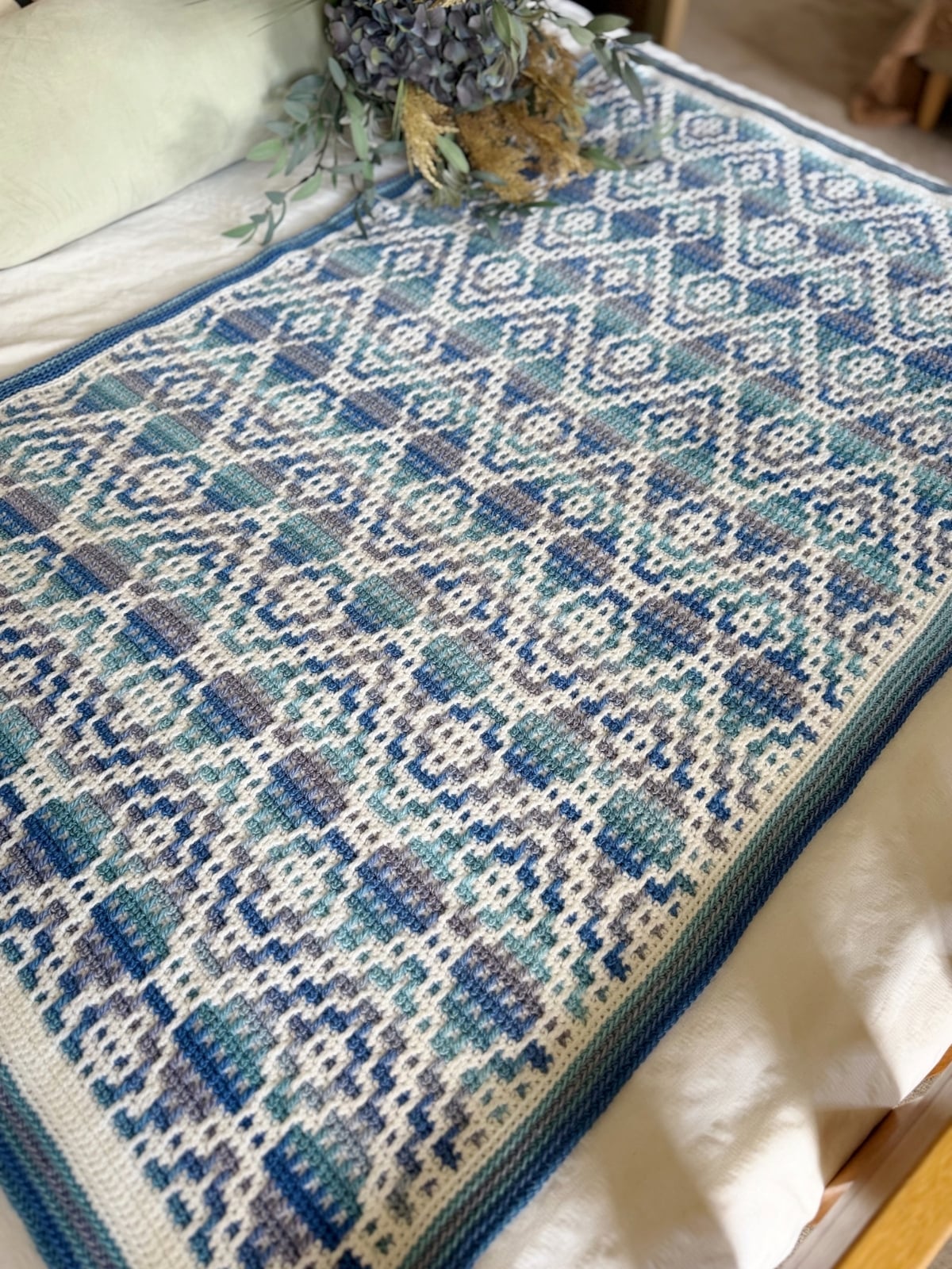 A blue and white patterned mosaic crochet blanket is laid flat on a bed, with a bouquet of dried flowers resting at the head of the bed.