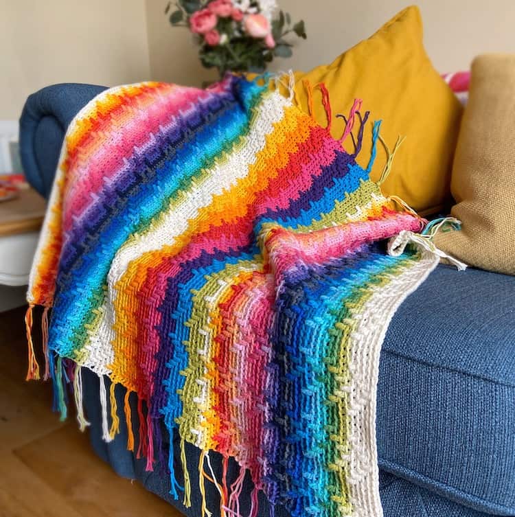 rainbow overlay mosaic crochet blanket pattern with tassels laid on a blue sofa with yellow cushions