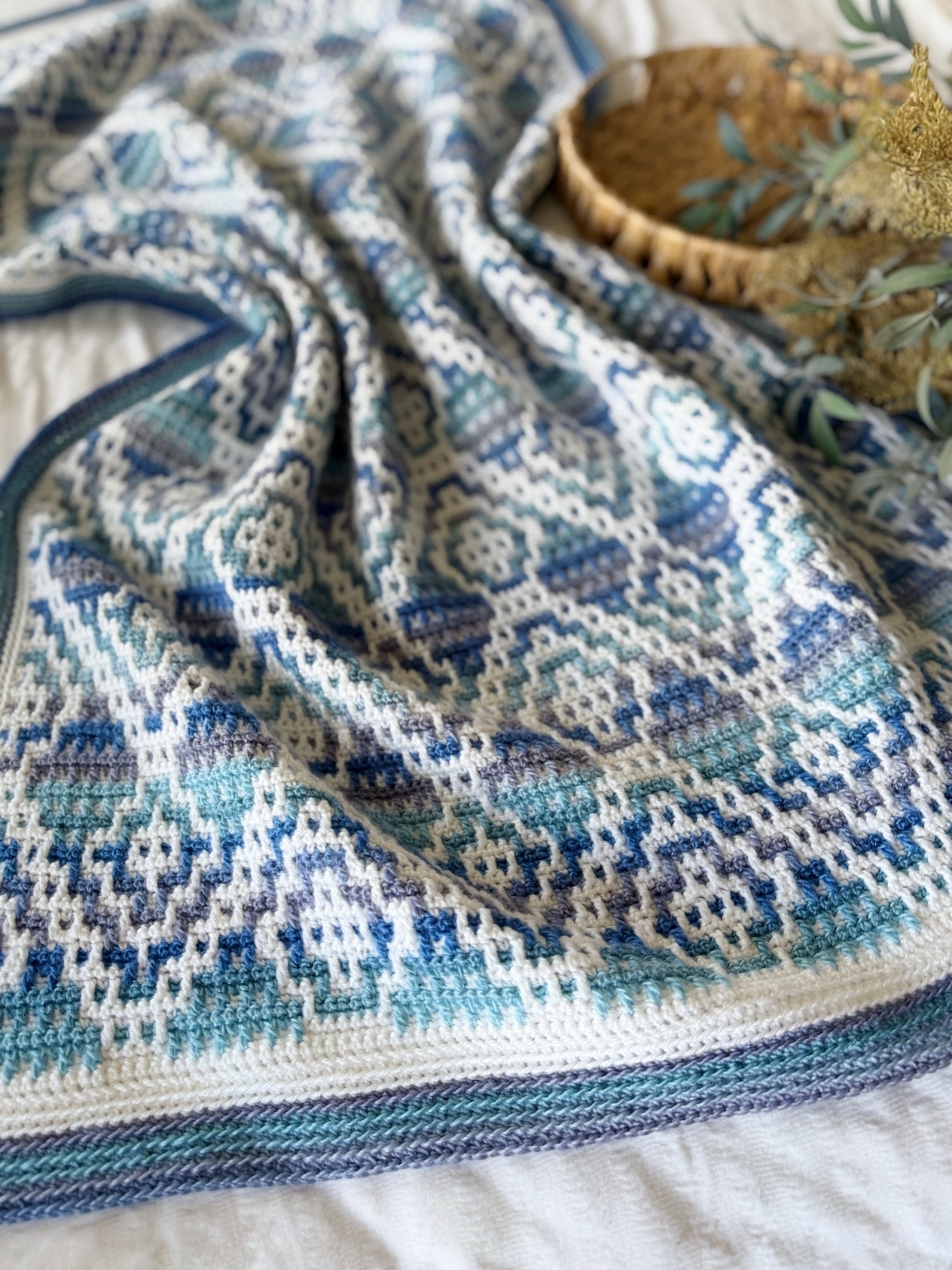 A blue, white, and teal mosaic crochet blanket with a geometric pattern is spread out on a bed next to a small wicker tray with greenery.
