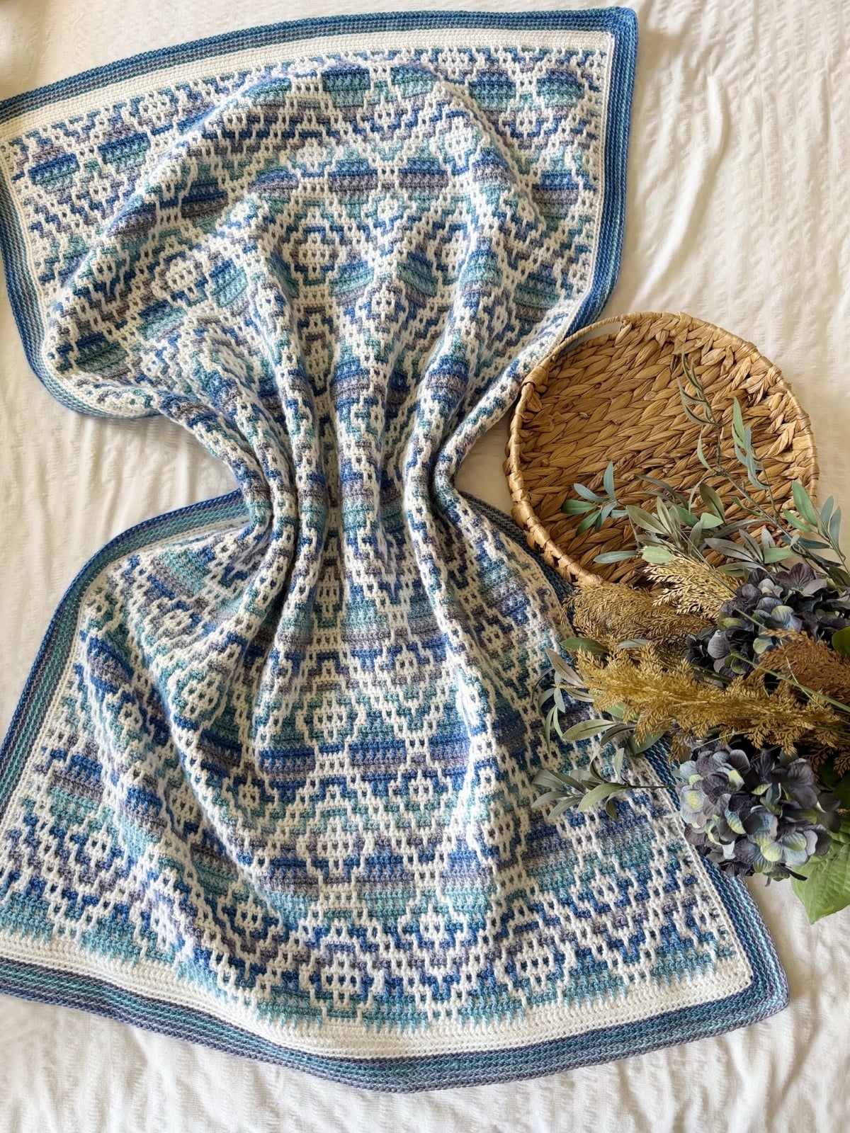 A mosaic crochet blanket with a blue and white geometric pattern is displayed on a bed next to a woven tray and a bundle of greenery and purple flowers.