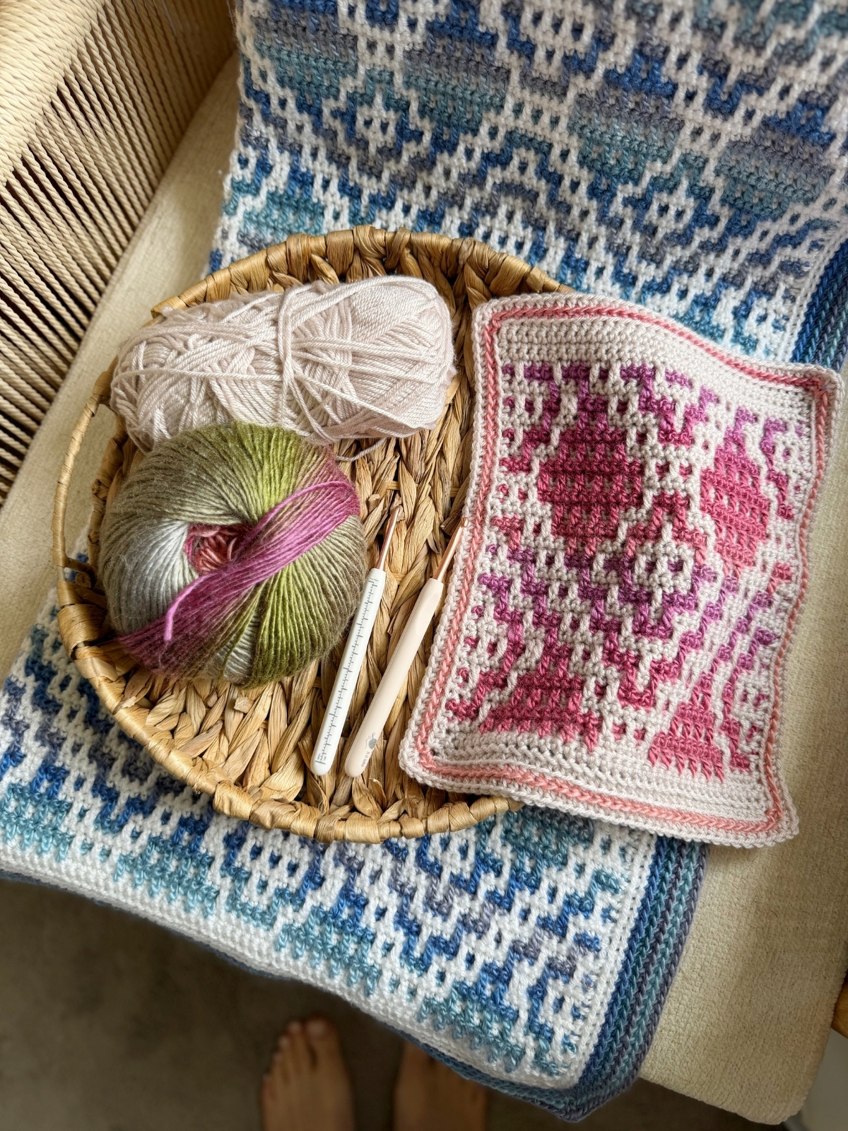 A woven basket with yarn, crochet hooks, and a pink and white crochet swatch sits on a blue mosaic crochet blanket draped over a chair.