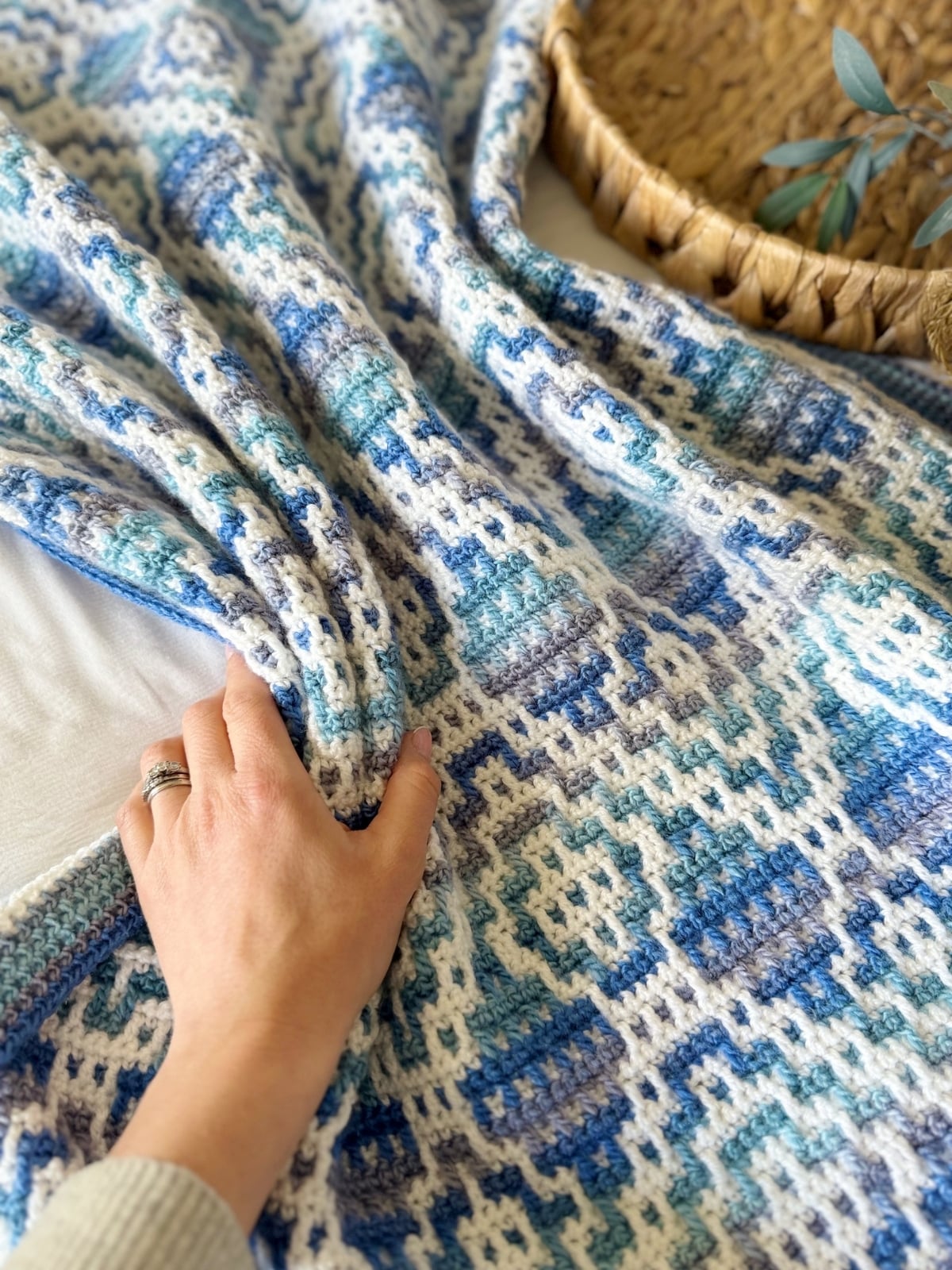 A hand rests on a blue and white patterned mosaic crochet blanket, with a woven basket and green foliage visible in the background.