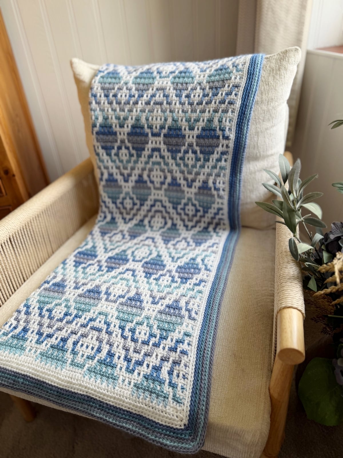 A mosaic crochet blanket with blue, white, and teal geometric patterns is draped over a beige cushion on a wooden chair.