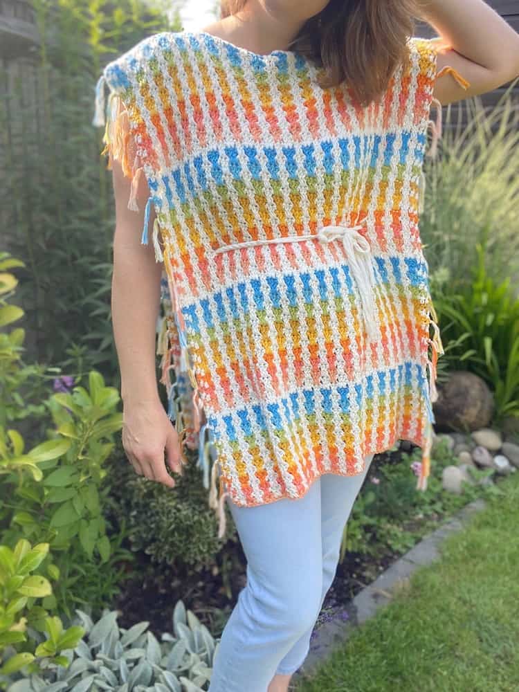 A person standing in a garden wears a colorful summer crochet poncho with a white tie belt, paired with light blue pants.