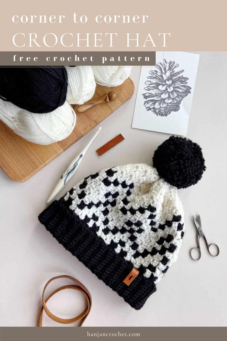 black and white crochet hat using corner to corner pattern with yarn, scissors, crochet hook surrounding it