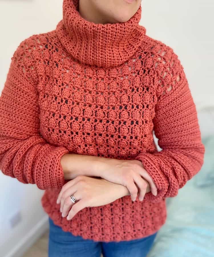 woman rolling sleeves up wearing bright coral crochet sweater with turtle neck