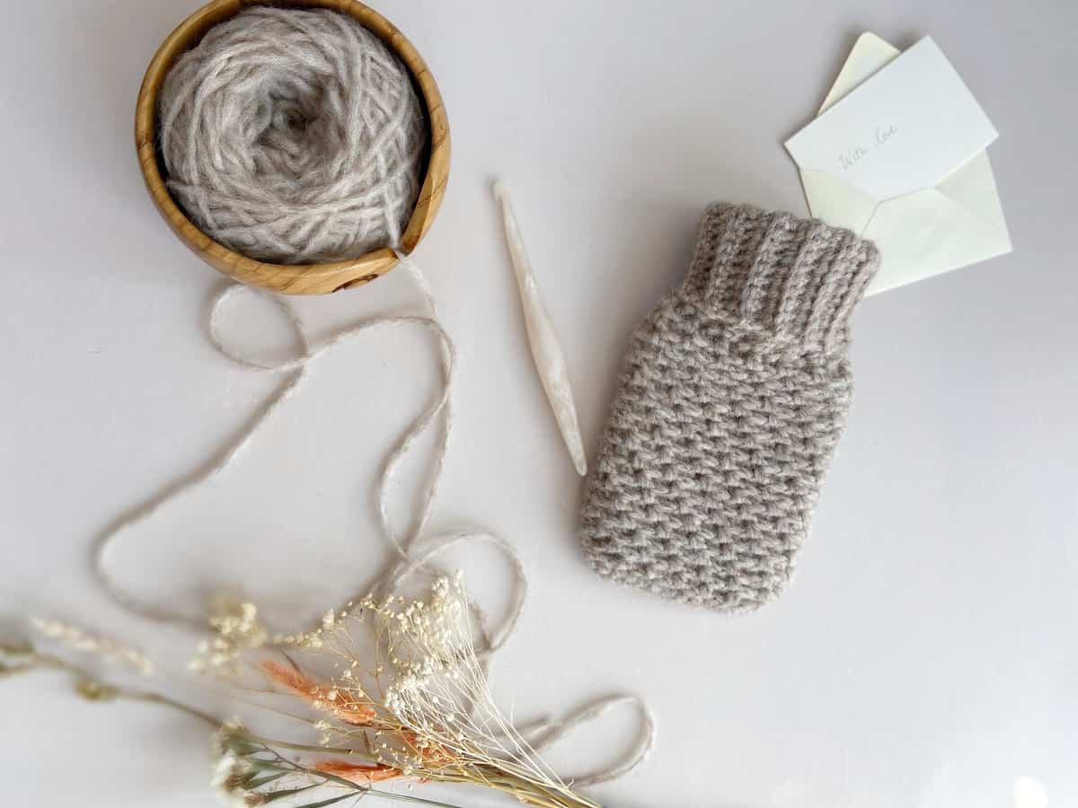 Fluffy, gray hot water bottle cover with yarn in wooden yarn bowl, flowers, card and crochet hook.
