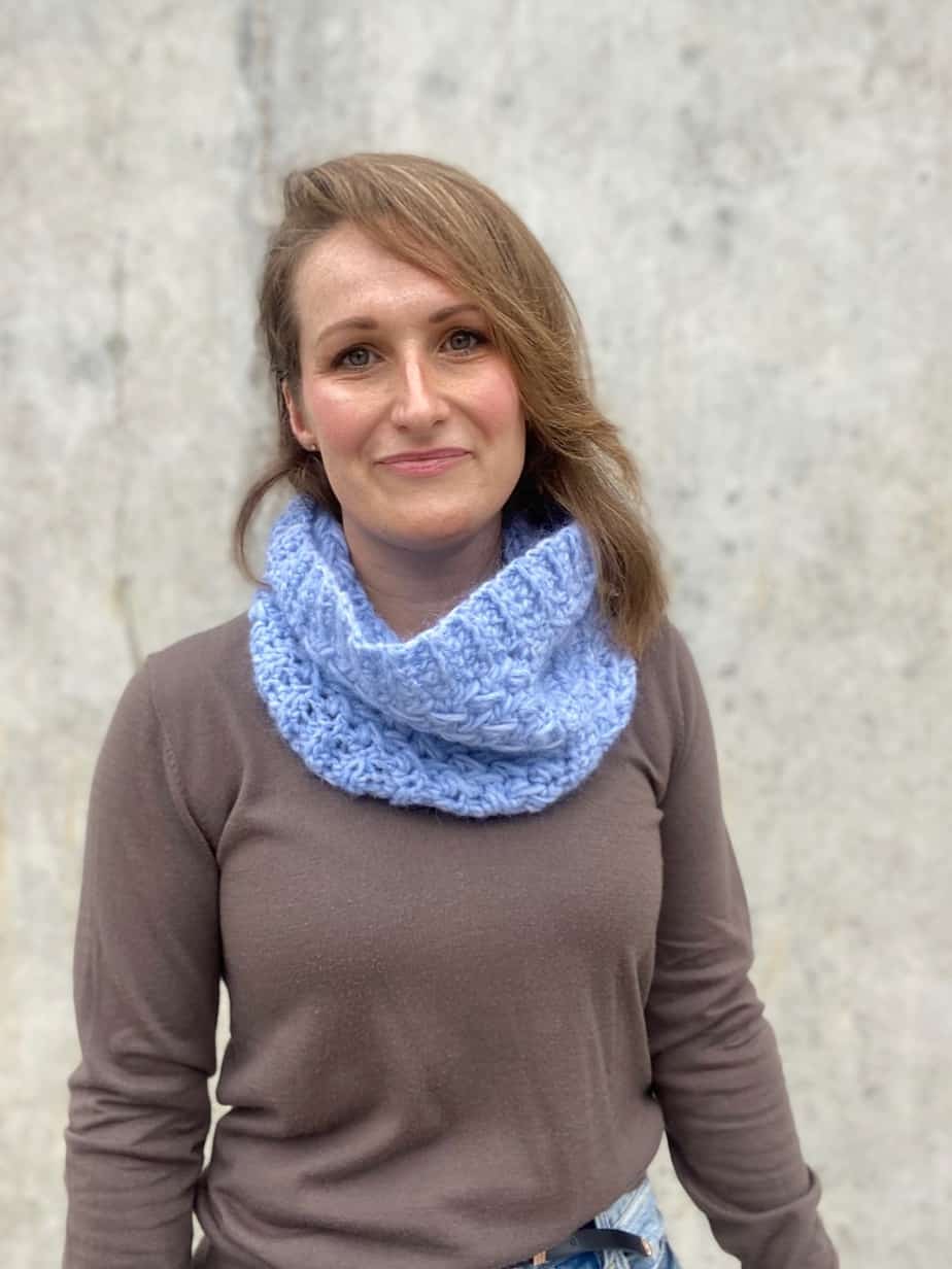 Woman looking at camera wearing fawn jumper and light blue crochet cowl.