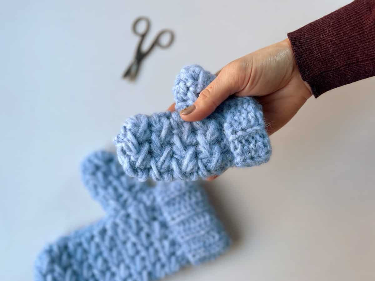 Someone holding a tiny feather stitch crochet mitten in their hand with scissors and larger mitten in the background.