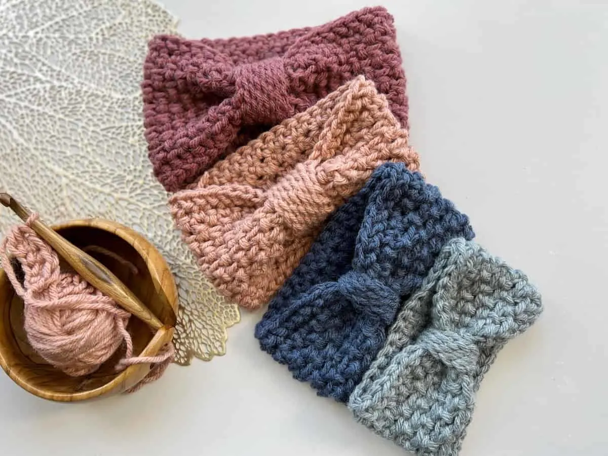 four handmade chunky crochet headbands with a crochet hook and yarn in yarn bowl on the side