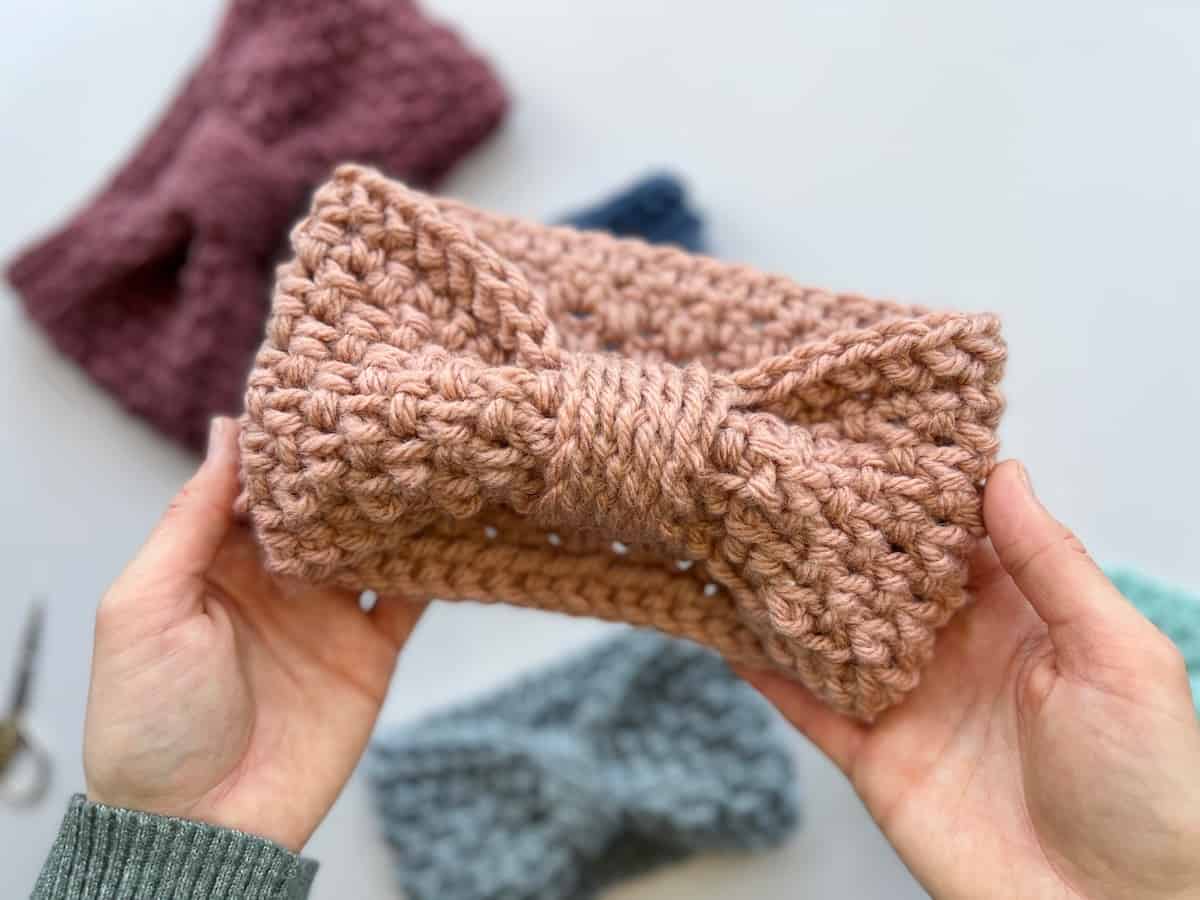 hands holding chunky crochet headband in peach colour with other headbands in background on a white table