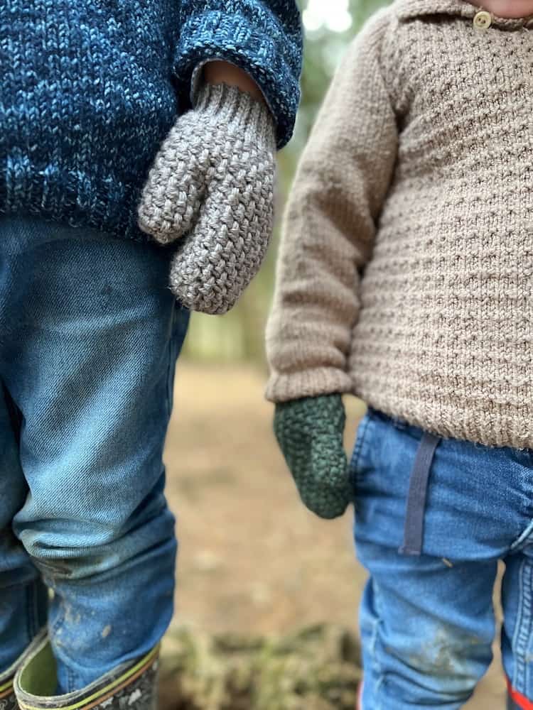 close up of two children with arms by sides wearing moss stitch crochet mittens, wellies and muddy jeans