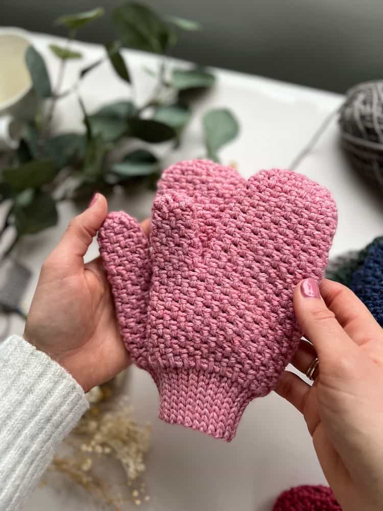 person holding pink moss stitch crochet mittens over a desk with yarn and flowers in background