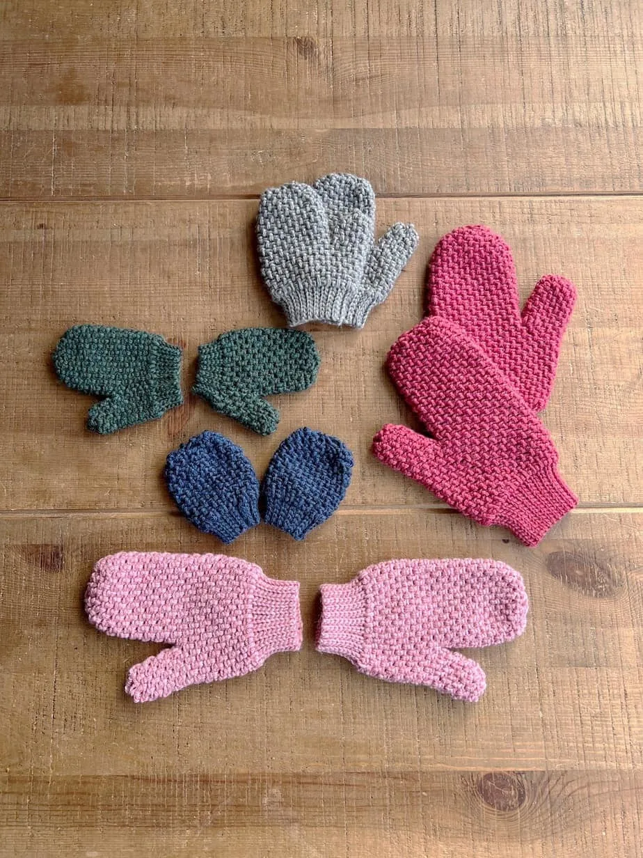 Five pair of crochet mittens from baby to adult size laid on dark wood table