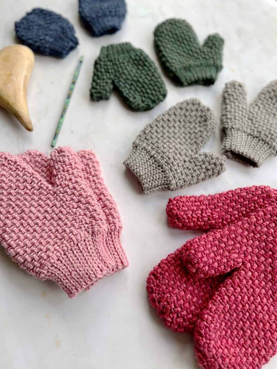 angled photo of four pairs of herringbone moss stitch crochet mittens on a table with crochet hook and wooden heart