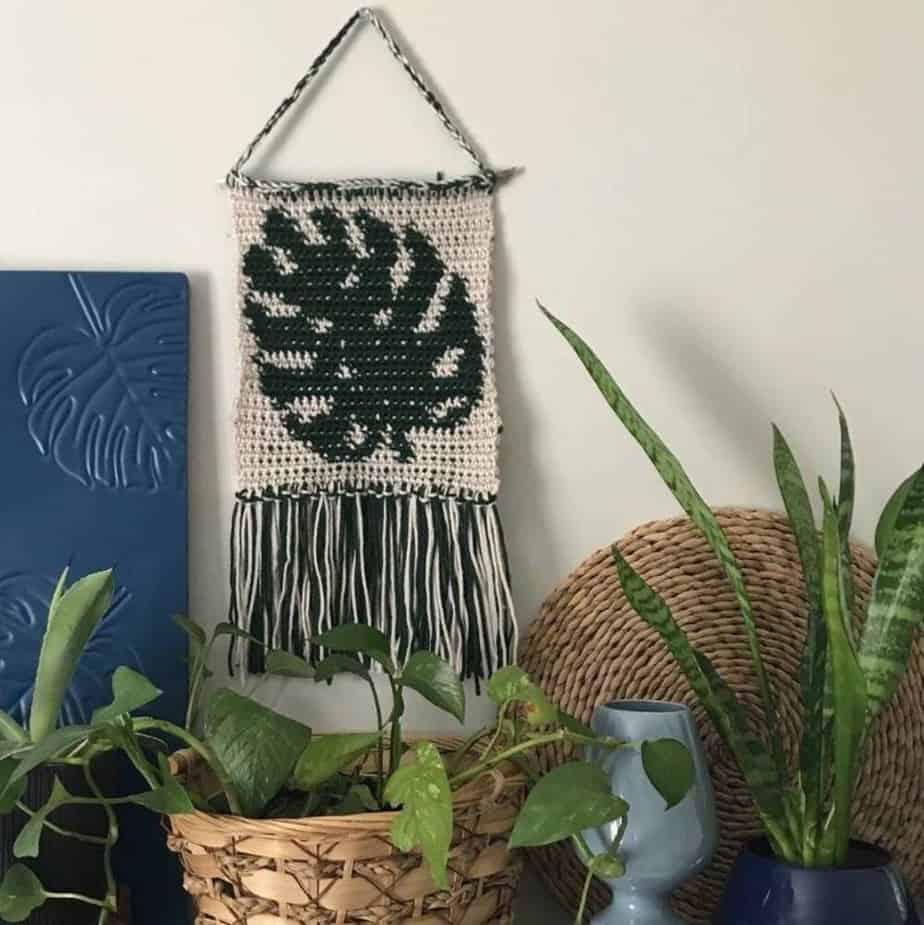 Crochet wall hanging with leaf design, hanging on wall with plants around it.