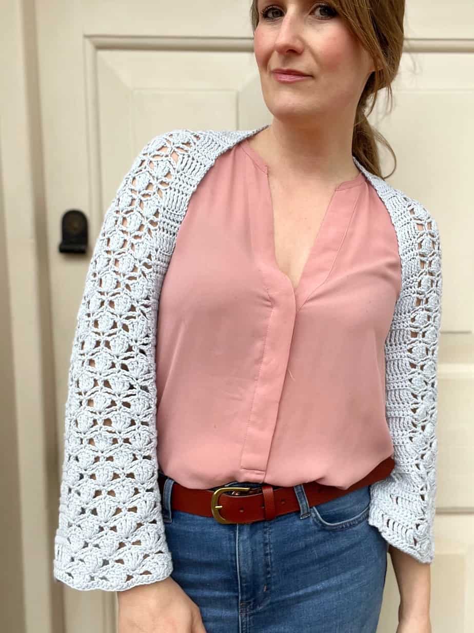 woman looking at camera wearing a pink top and light blue lace crochet shrug with crochet shell stitch detail