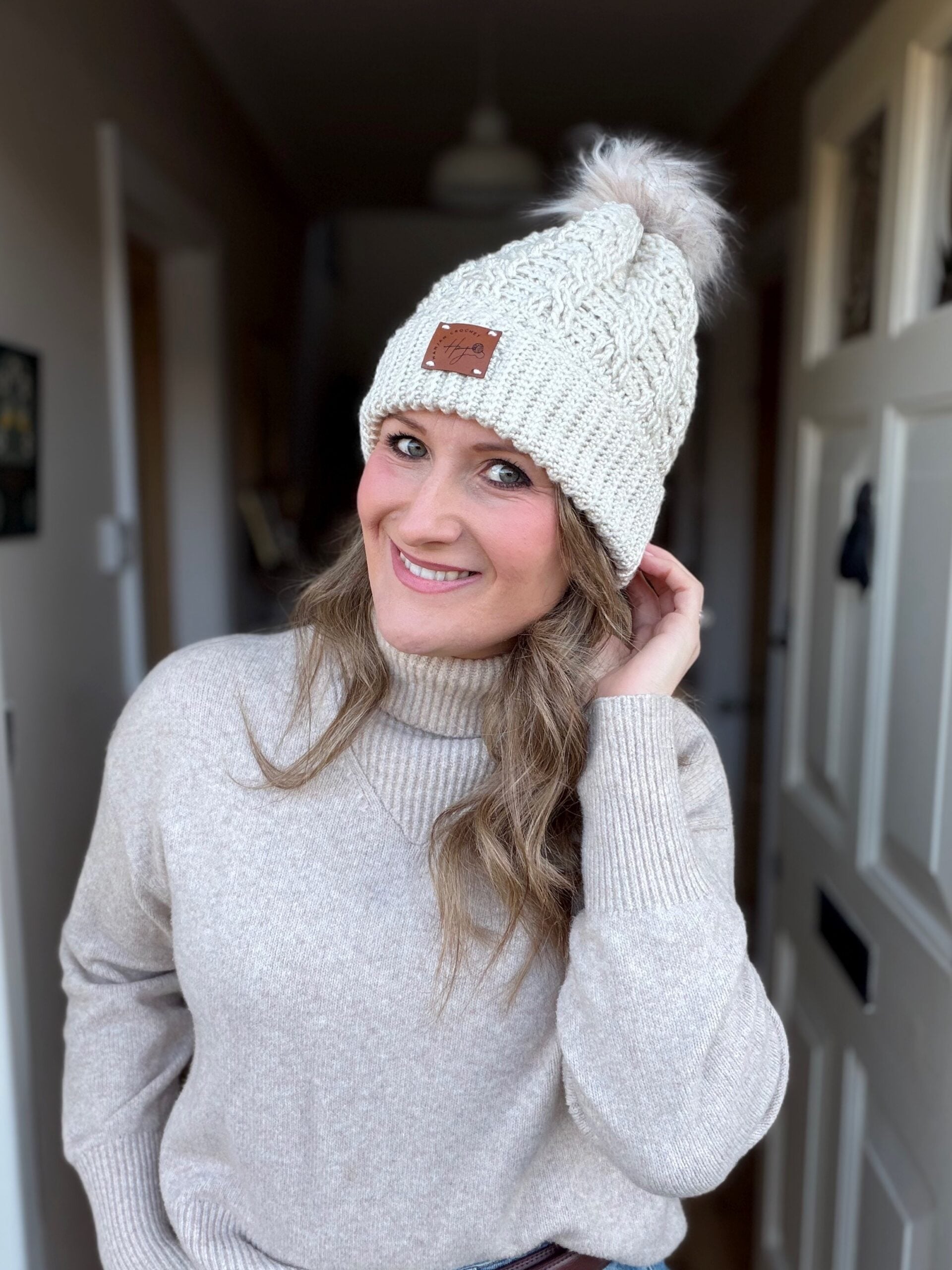 Woman in a beige sweater and a quick crochet hat with a pom-pom stands indoors, smiling and touching her hair.