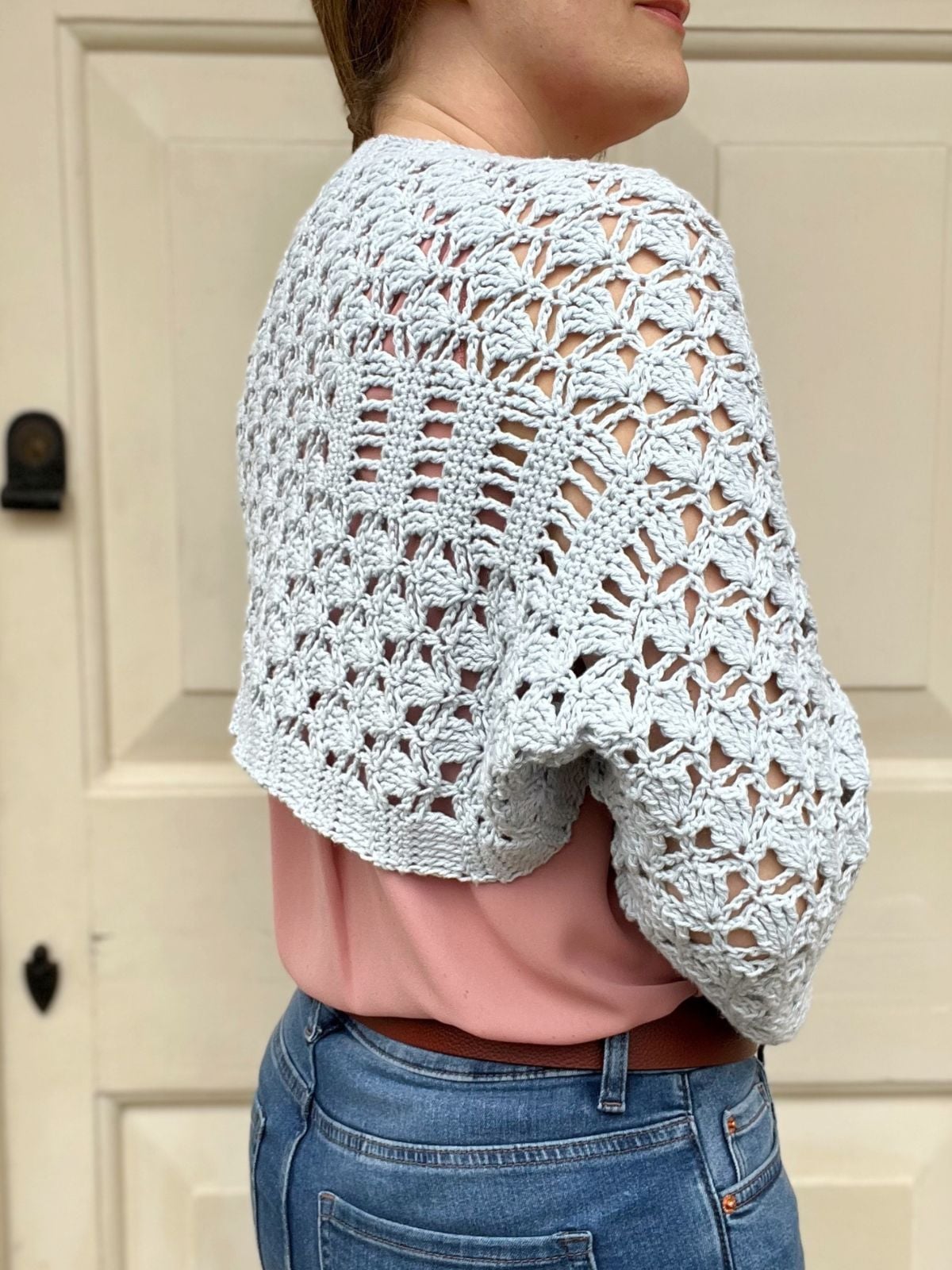 A person wearing a light blue, lacy crochet wrap pattern shawl over a pink top and blue jeans, standing in front of a beige door.