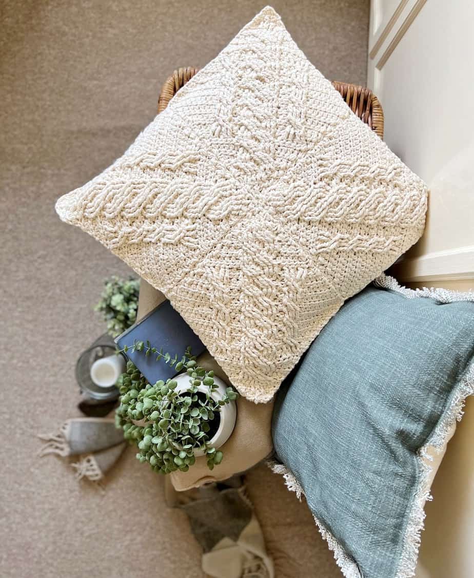 cream cable crochet pillow sitting on large basket with plants books and blanket