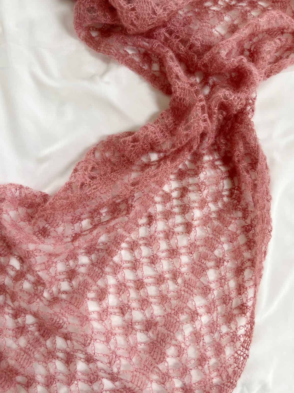 A pink, lacy mohair scarf with an intricate openwork crochet pattern is draped on a white surface.