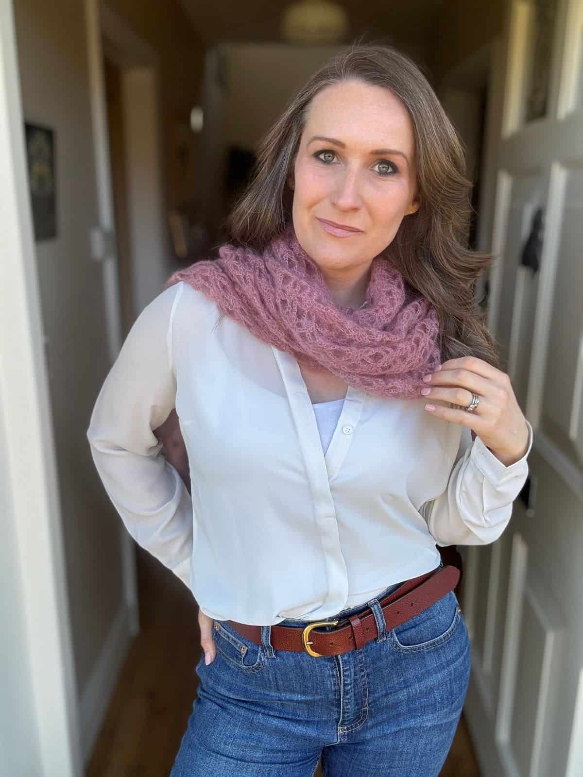 A woman in a white shirt and blue jeans stands indoors, her look accentuated by a pink mohair crochet scarf pattern and a brown belt, with one hand confidently on her hip.