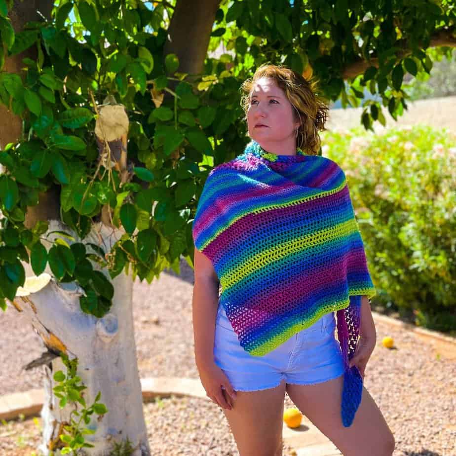 Woman in shorts under a tree wearing rainbow crochet shawl.