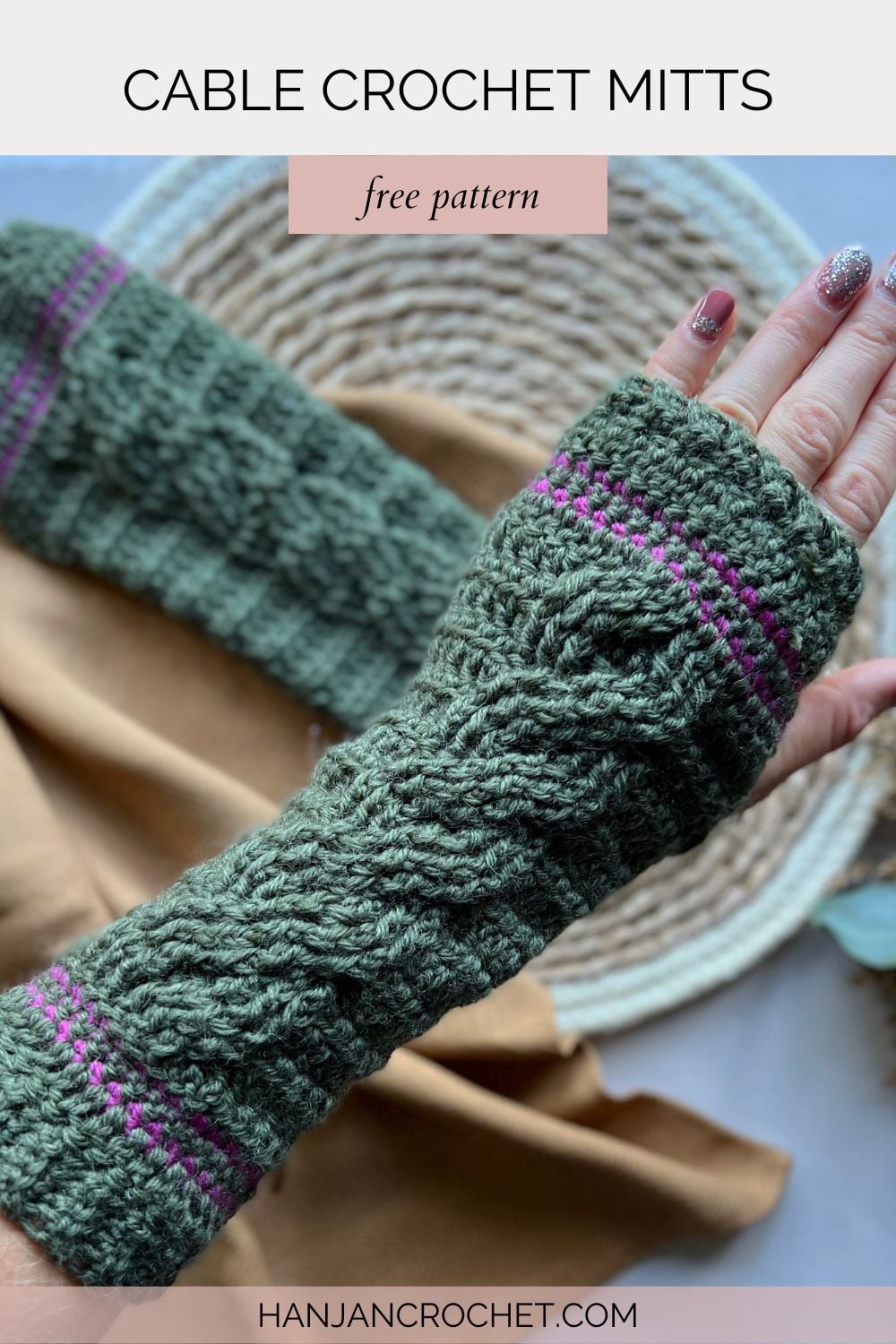 A hand wearing green crocheted mitts with pink stripes, crafted using a cable crochet wrist warmer pattern, is displayed on a beige background next to a woven mat.