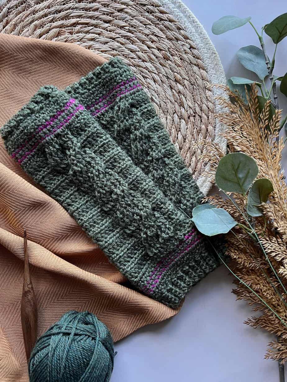 Crochet cable wrist warmers laid on table with crochet hook and ball of yarn.