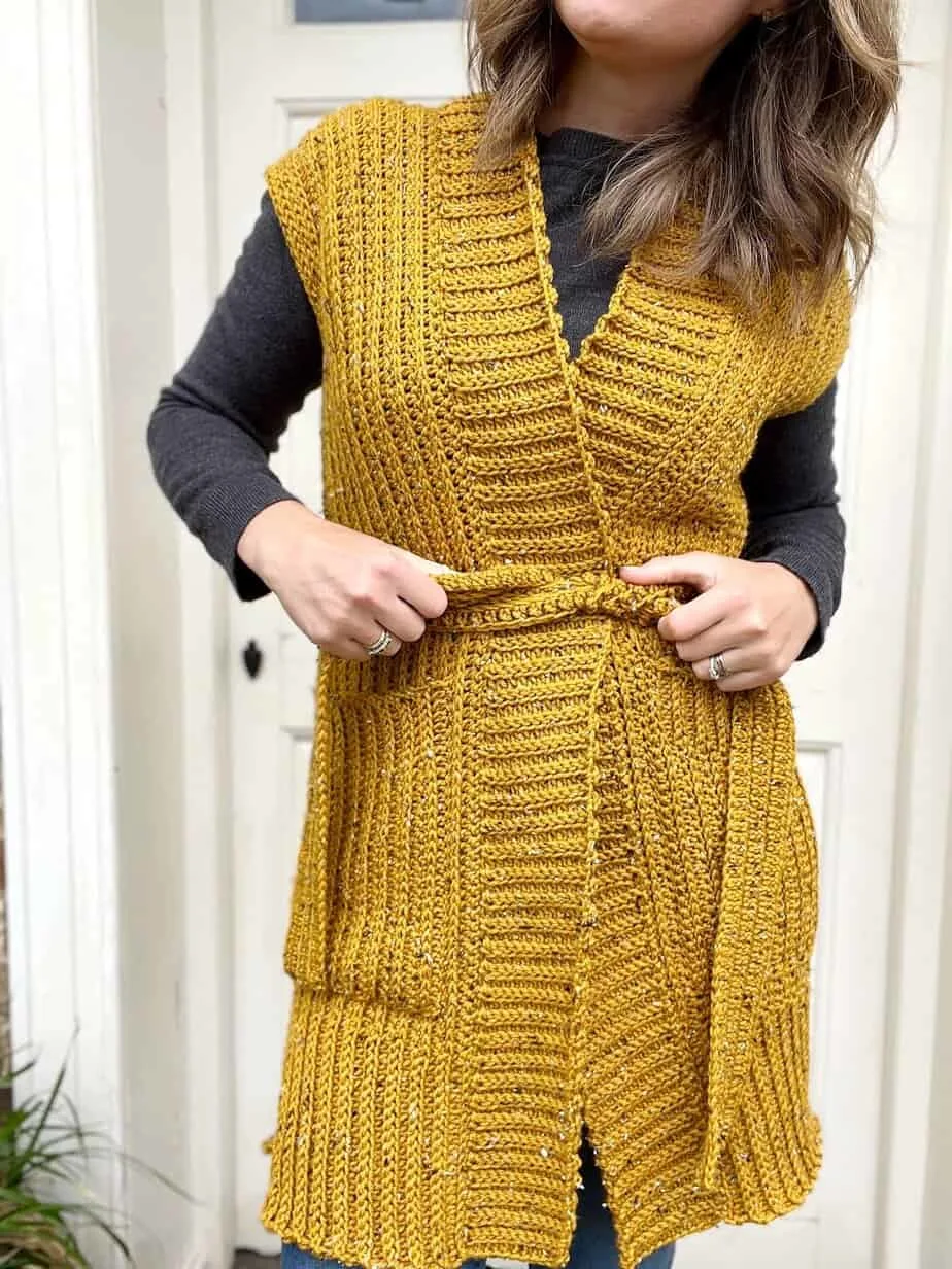 Woman wearing long sleeveless crochet cardigan with pockets and belt.
