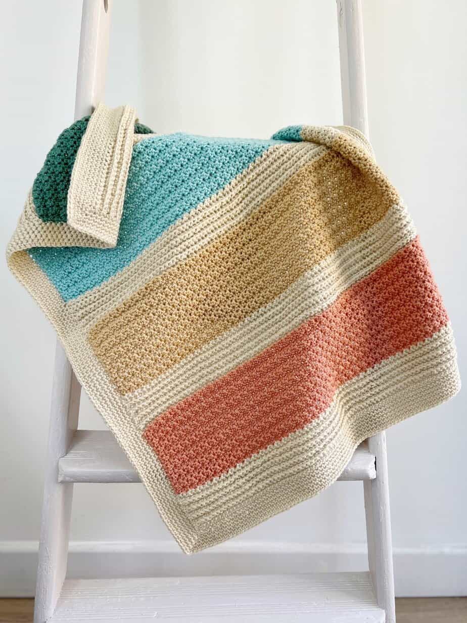 Striped baby crochet blanket pattern in coral, blue and green on a ladder.