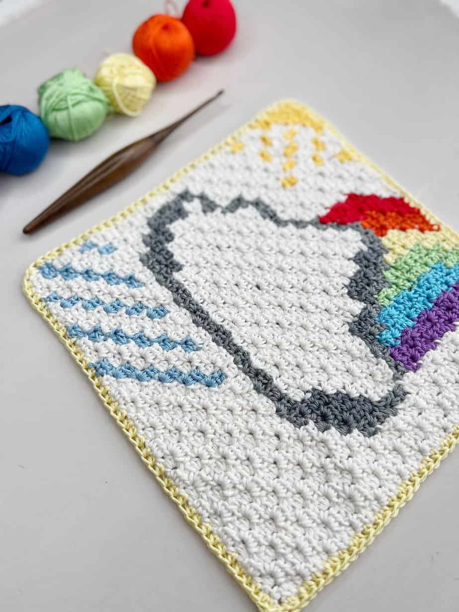 Close up of C2C square pattern of cloud and rainbow with crochet hook and yarn.
