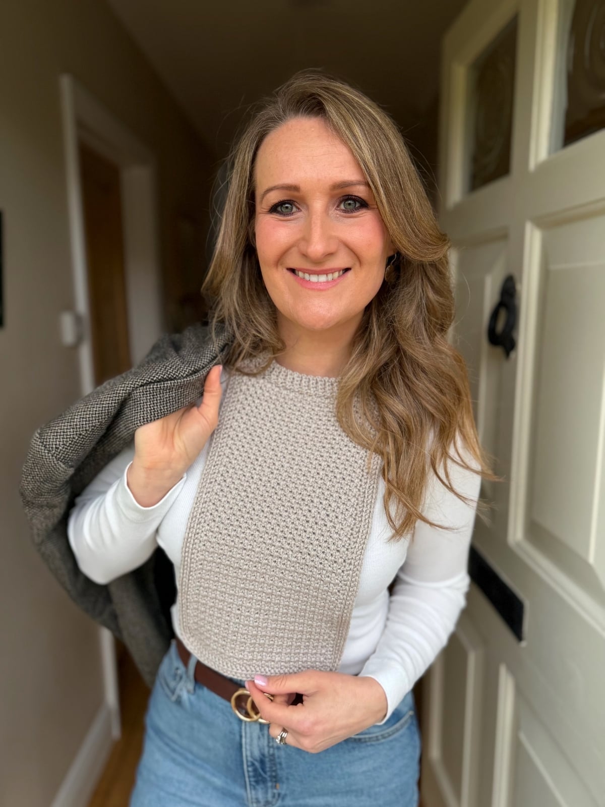 Woman with long blonde hair wears a white long-sleeve top and a textured beige bib, reminiscent of a crochet dickie pattern, holding a gray blazer over her shoulder while standing in a hallway near a white door.
