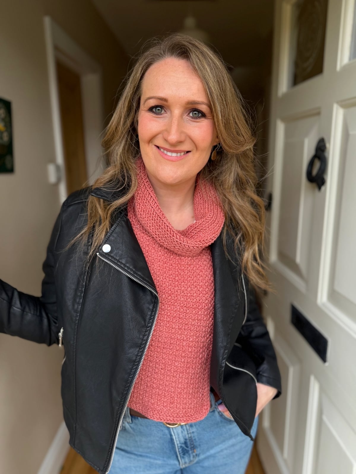 A woman with long, wavy hair wearing a black leather jacket, pink crochet dickie pattern sweater, and blue jeans stands indoors near a white door, smiling at the camera.