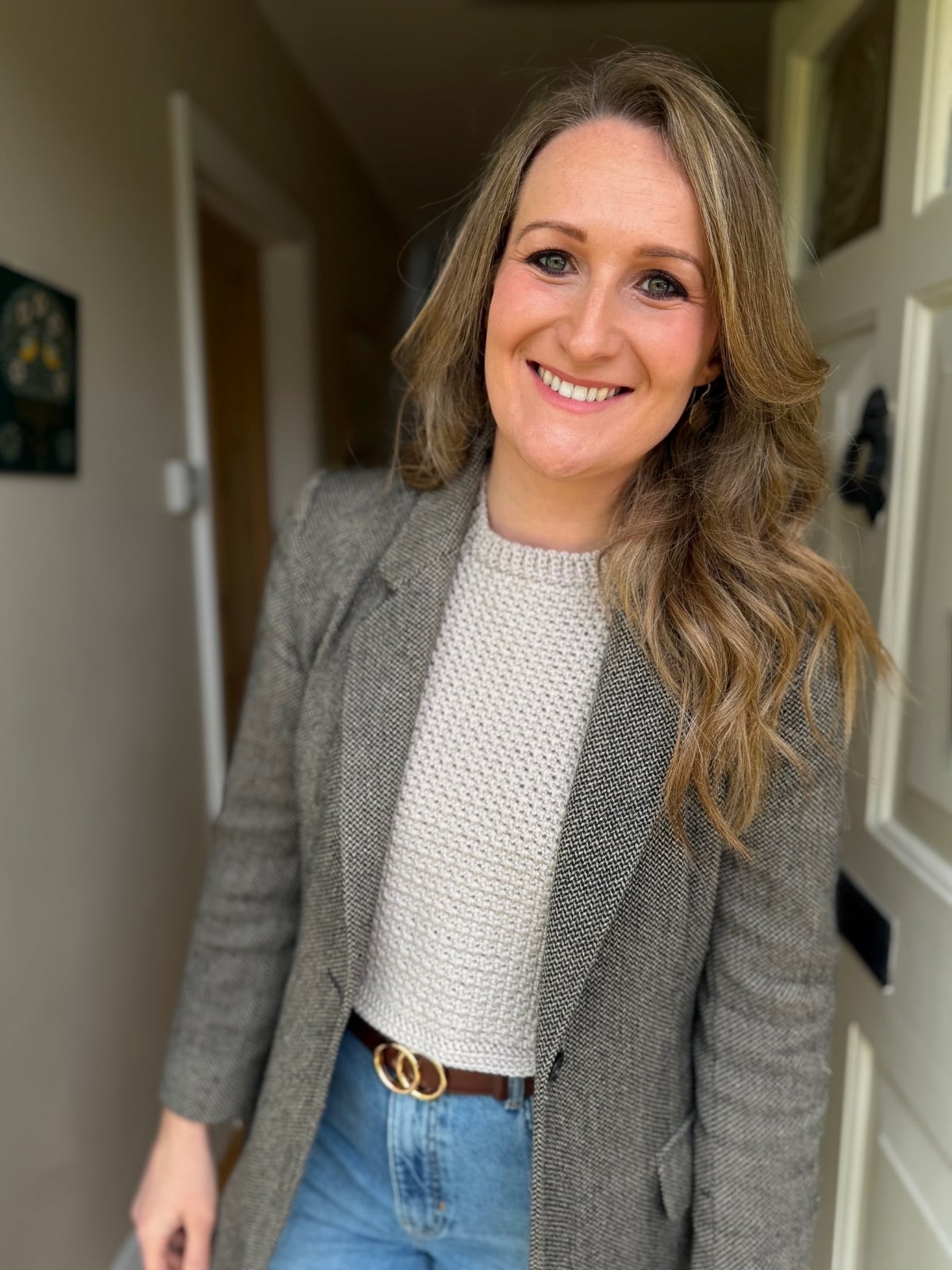 A woman with wavy blonde hair wearing a grey blazer, beige sweater layered with a crochet dickie pattern, and blue jeans stands indoors, smiling at the camera.