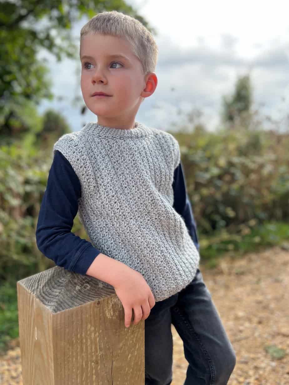 Young boy leaning on post wearing crochet vest pattern in grey.