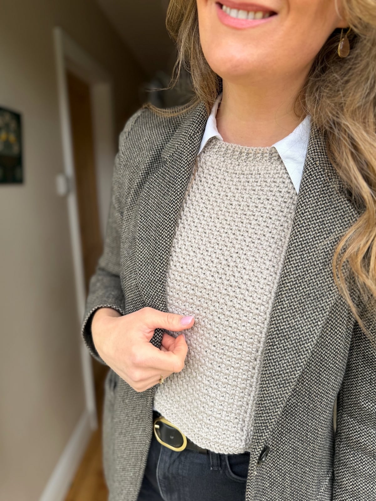 A person wearing a grey textured blazer, beige knit sweater styled with a crochet dickie pattern, and black pants with a belt, standing indoors and smiling.