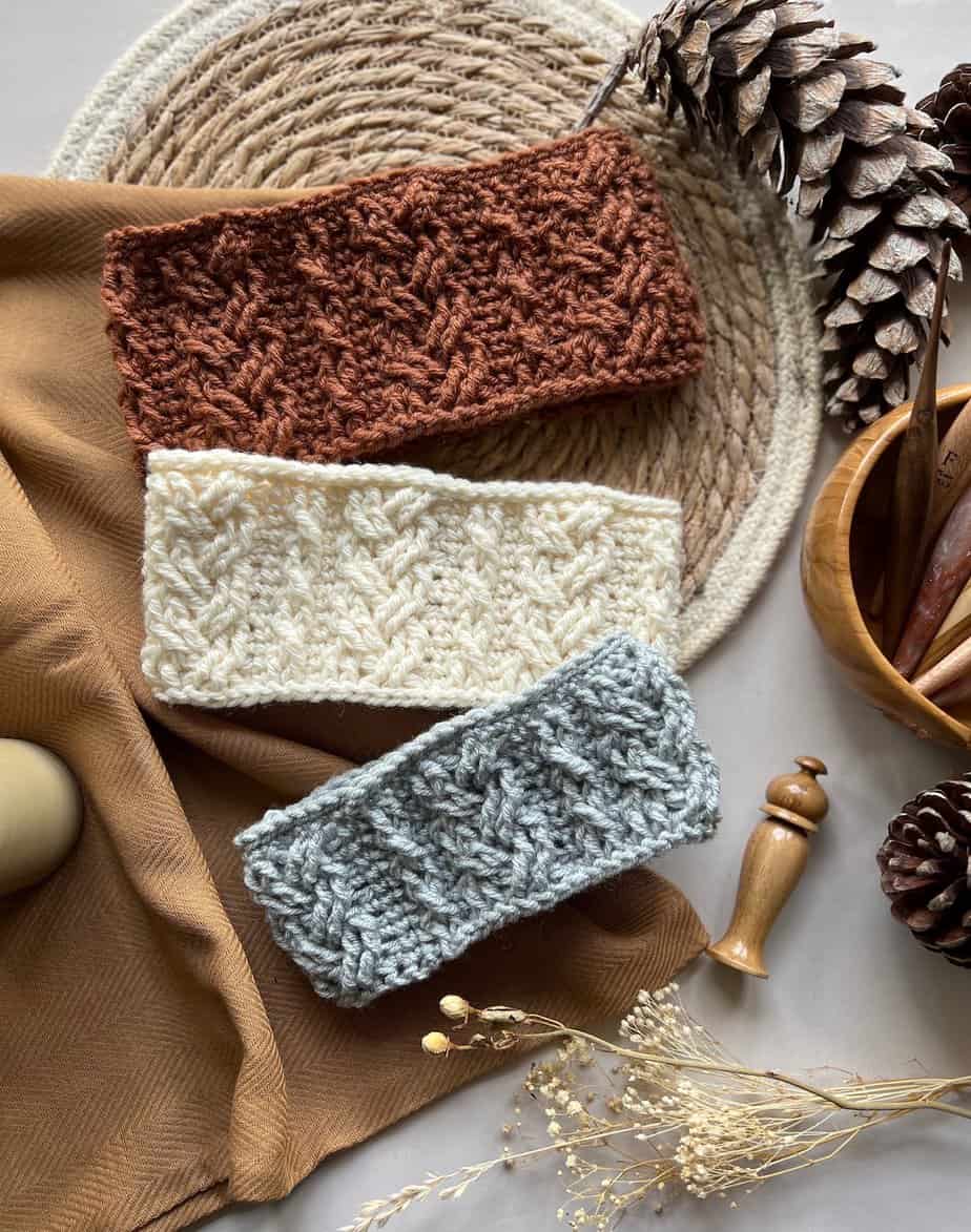 Three crochet headbands with cable stitches laid on table with crochet hooks and pine cones.