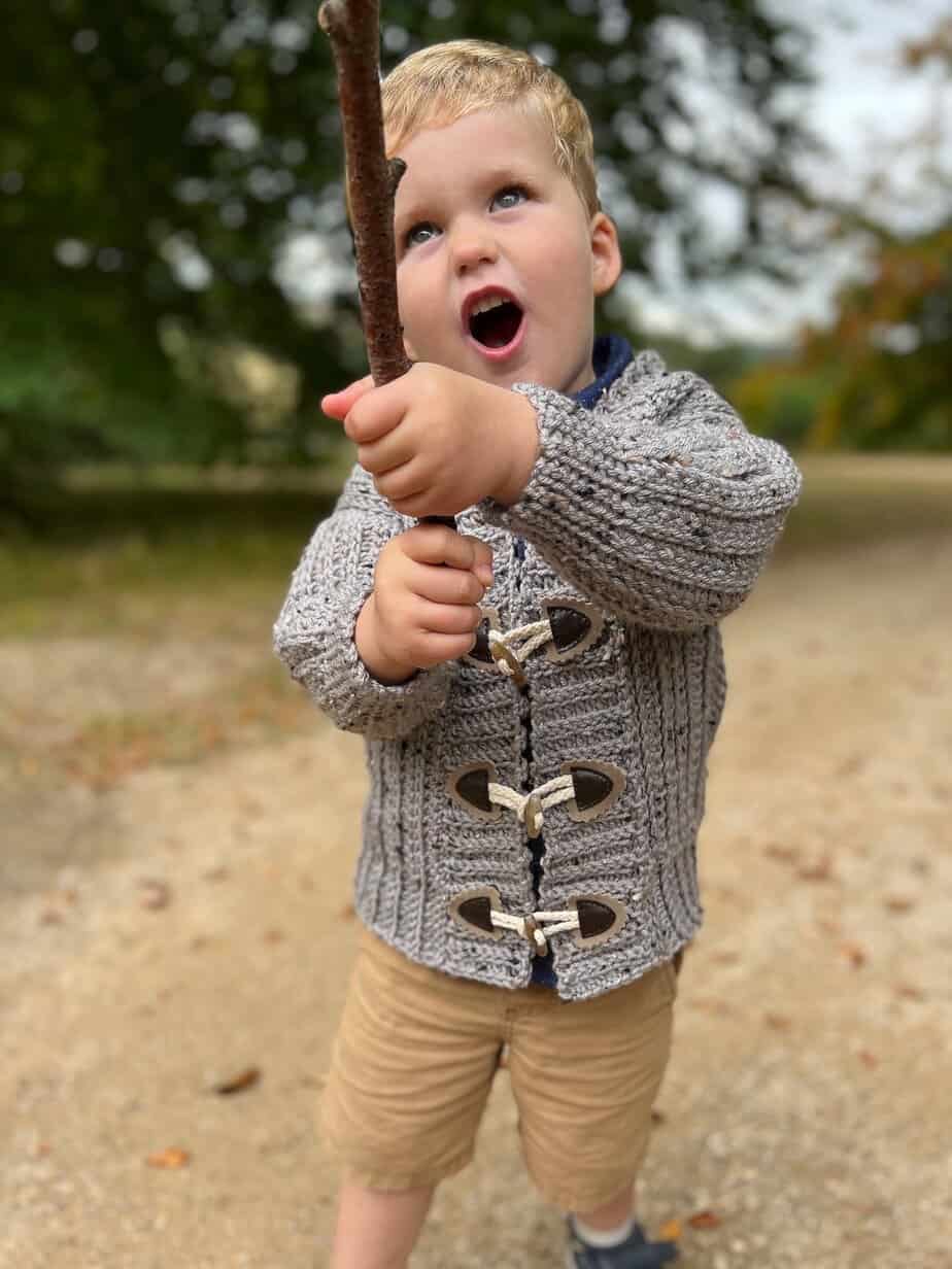 Boy holding a stick wearing easy crochet cardi with toggles.