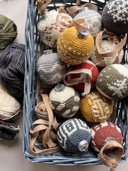 A basket filled with crochet Christmas ornaments in various colors and patterns, including festive trees and fair isle crochet bauble patterns, surrounded by balls of yarn and ribbon.
