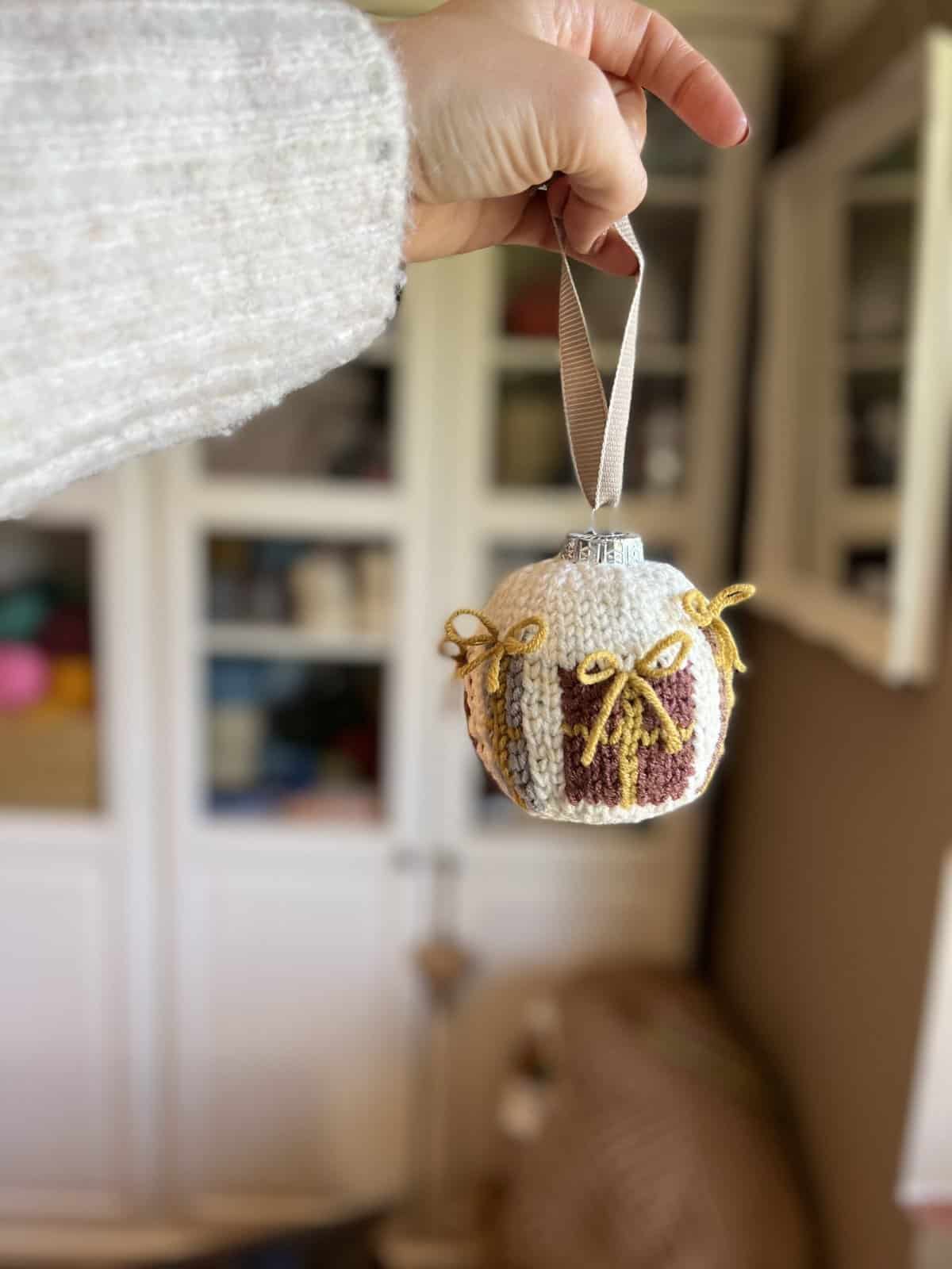 A hand holds a crochet Christmas present ornament, covered in knitted fabric with a brown design and small yellow bows, against a blurred indoor background.