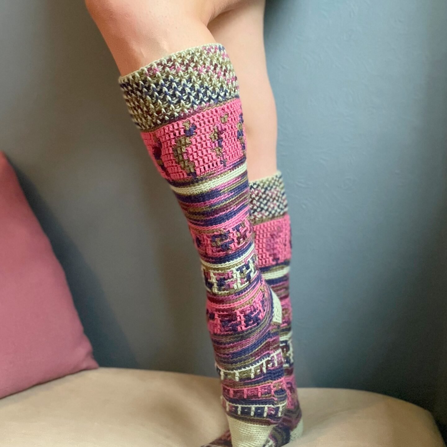 Knee high crochet socks with a gray background and a pink pillow nearby.