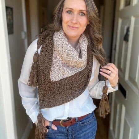 A person wearing a white blouse, blue jeans, and a brown scarf with tassels from an easy triangle shawl crochet pattern stands indoors.