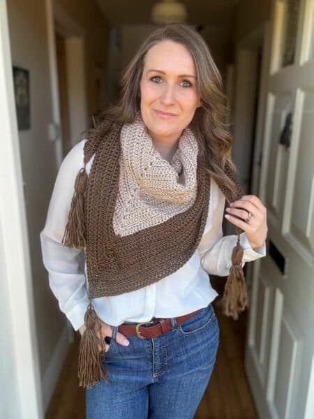 A person wearing a white blouse, blue jeans, and a brown scarf with tassels from an easy triangle shawl crochet pattern stands indoors.