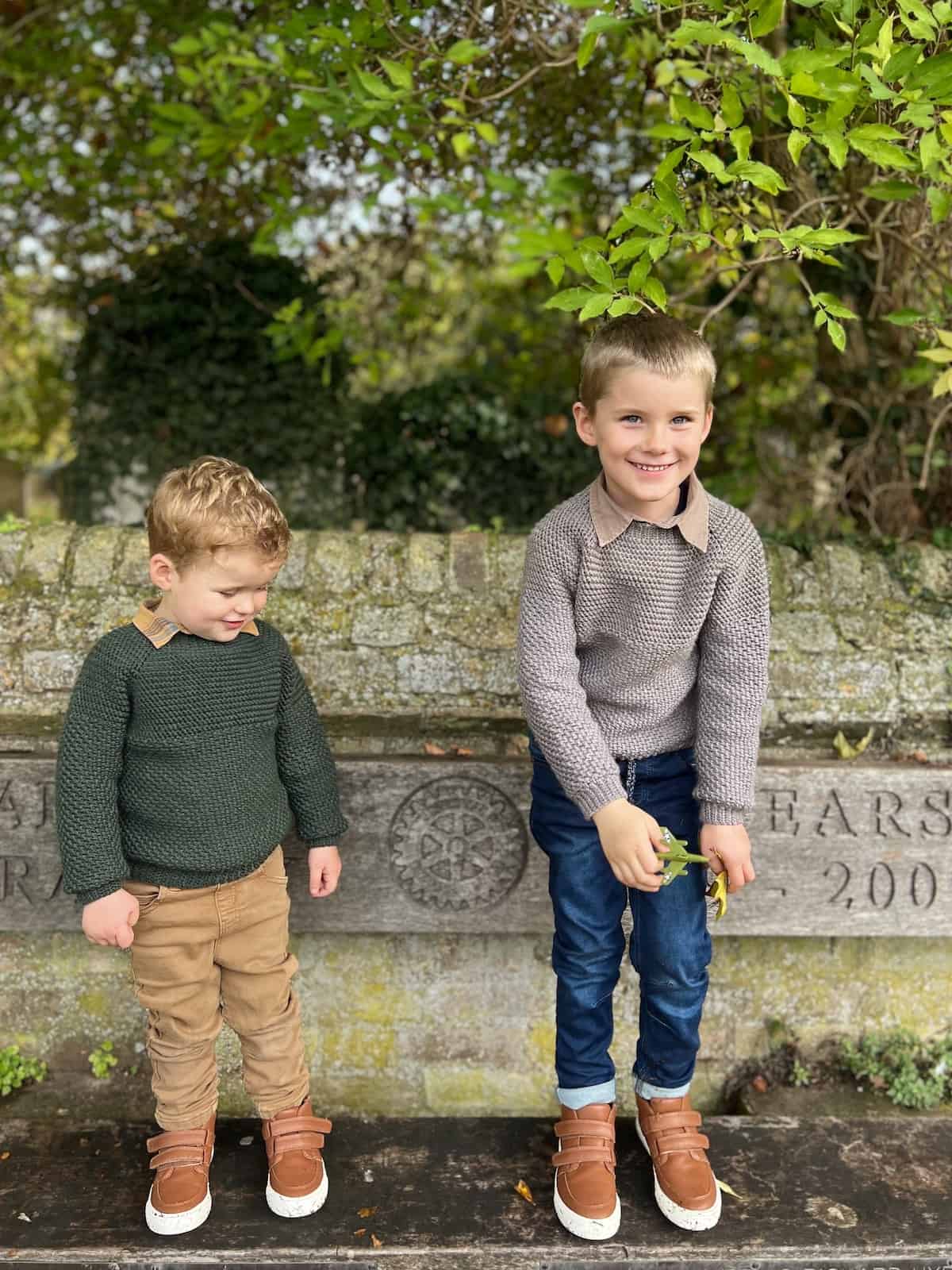 Two boys in crochet sweaters standing on a bench.