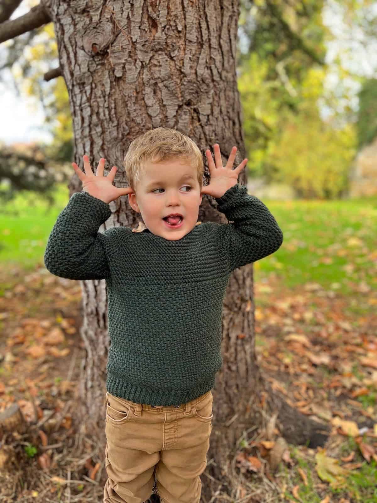 Toddler in crochet sweater with hands on ears and tongue sticking out.